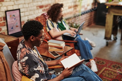 African American guy reading book while sitting in armchair in living room