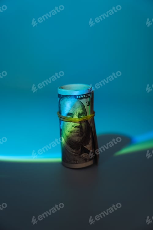 Preview: One hundred dollar bills rolled into a tube and wrapped with an elastic band under hard blue light