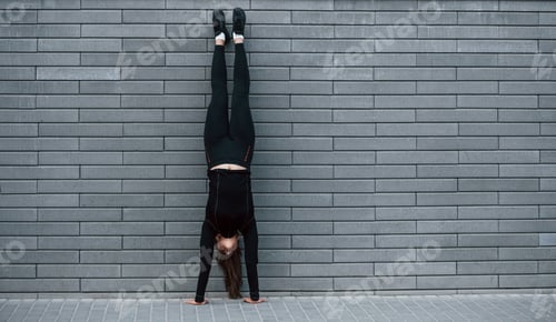 Preview: Young sportive girl in black sportswear doing hard handstand exercises outdoors near gray wall