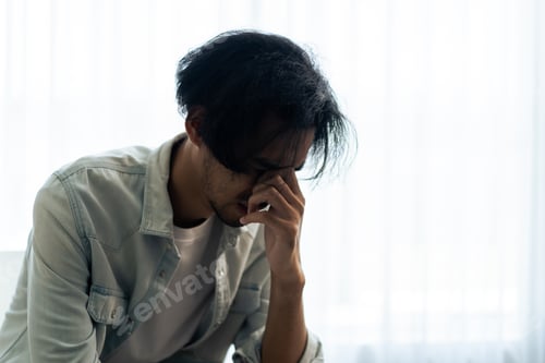 Preview: Asian young upset depressed man sitting alone in living room at home.