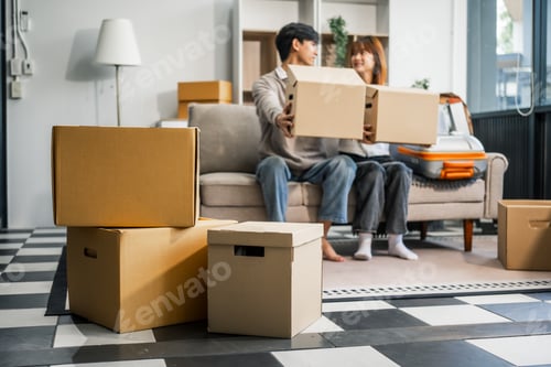 Preview: A young couple happily sits on sofa, packing their belongings into a big box for their move to