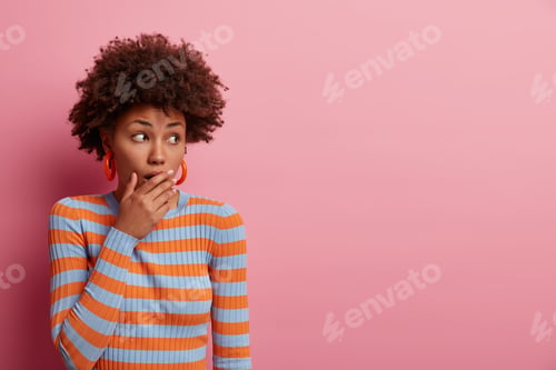 Preview: Concerned nervous young woman with fluffy curly hair, gasps from fear, covers mouth with hand, stand