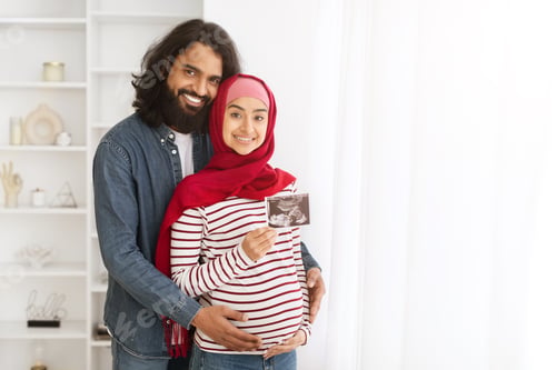 Preview: Pregnant Muslim Couple Holding Baby Ultrasound Scan While Standing In Home Interior