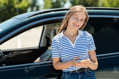 Preview: Young smiling woman using smartphone leaning on car door smiling