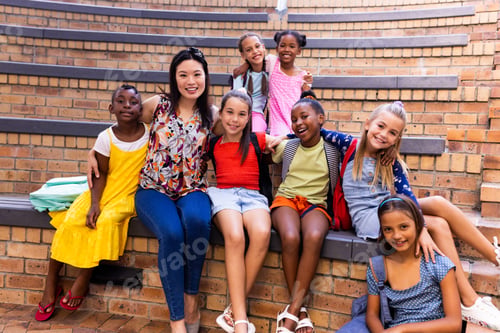 Preview: Portrait of diverse female teacher and schoolgirls sitting in elementary school outdoor auditorium