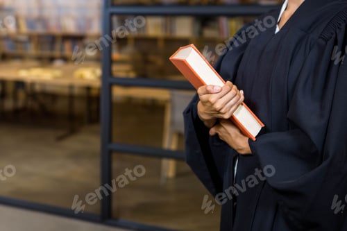 Preview: Female student wearing graduation gown standing by glass partition holding orange book in library