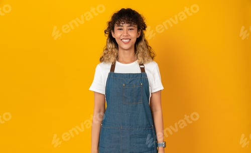 Preview: Studio shot of cheerful beautiful Asian woman wear apron and stand with smile on orange background.