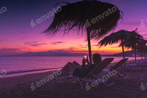 Preview: A woman photographing the sunset sitting on a relaxing beach chair with a parasol at night