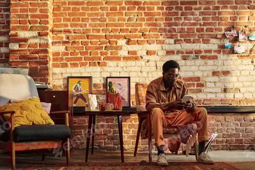 Preview: Young relaxed black man in casualwear sitting in armchair by brick wall