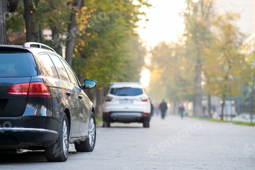 Preview: Cars parked in a row on a city street side on bright autumn day with blurred people walking on
