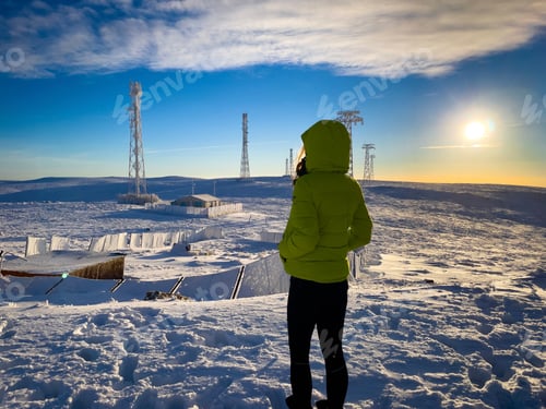 Preview: Rear view of young woman wearing green coat looking at transmission towers in the distance at sunset