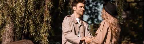 Preview: panoramic crop of man in trench coat looking at woman in hat while holding hands