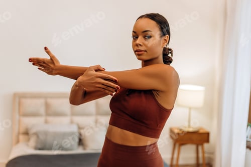 Preview: Fit young African American woman stretching arm at home, looking at camera in bedroom at home