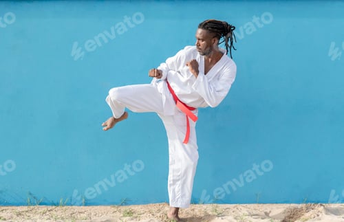 Preview: a young african american man training taekwondo outdoors kicking on a blue background