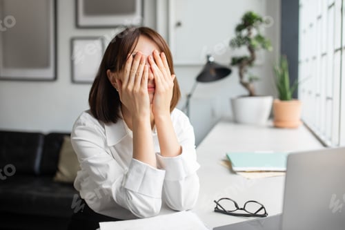 Preview: Tired sad young businesswoman covering face with hands, suffering from overwork, sitting at table