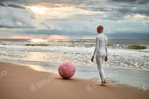 Preview: Hairless performer girl with alopecia in futuristic suit walking along seacoast with pink sphere