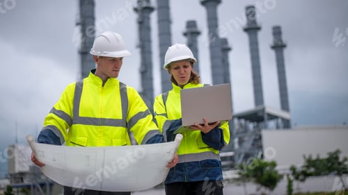 Preview: Engineers wearing safety gear, including hard hats examining survey a large blueprint tablet standin