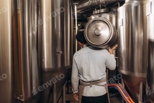 Preview: Shot of young man with metal beer barrels at brewery. Brewer carrying keg at brewery plant.