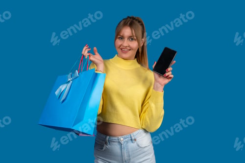 Preview: Young woman showing smartphone and holding shopping bag on blue background
