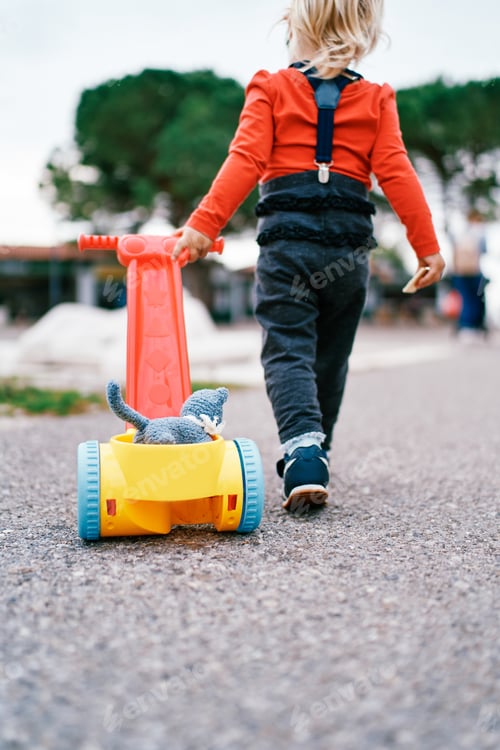Preview: Little girl is carrying a soft toy in a toy cart on asphalt. Back view