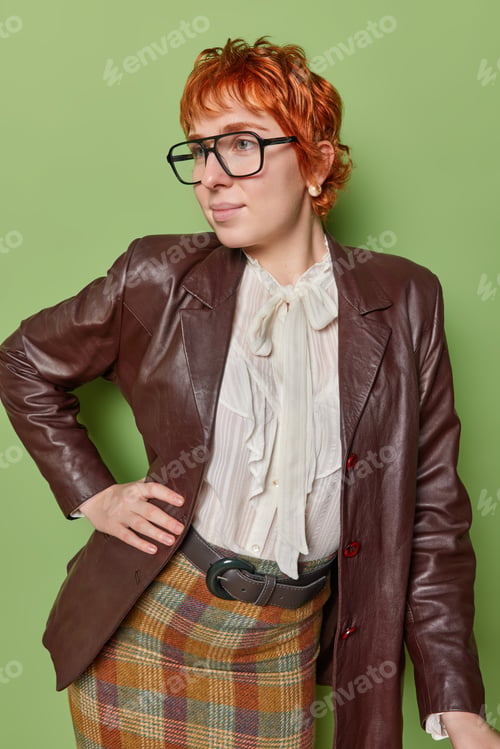 Preview: Indoor shot of thoughtful redhead lady dressed in elegant clothes keeps hand on waist focused aside