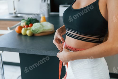 Preview: Measures waist. Young european woman is indoors at kitchen indoors with healthy food