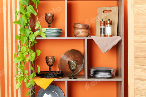 Preview: Shelving unit with different dishware and glasses near orange wall indoors