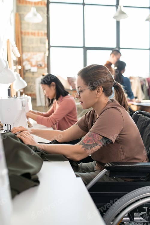Preview: Young disable seamstress sitting in wheelchair by workplace in front of camera and sewing clothes