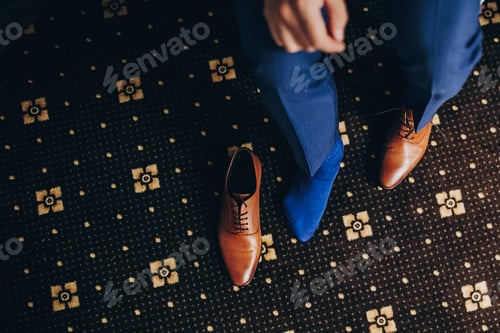 Vista previa: Elegante novio con traje azul que se pone zapatos marrones cerca de la ventana de la habitación del hotel