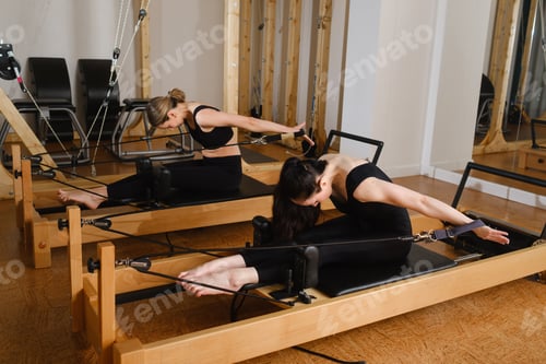 Preview: Two women in black sportswear are doing pilates on a reformer machine in the gym