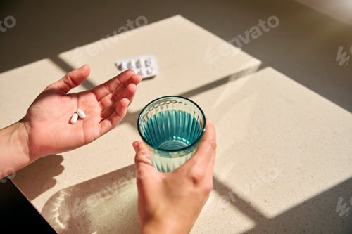 Preview: Close Up Looking Down On Woman Taking Medication At Home With Glass Of Water In Patch Of Sunlight