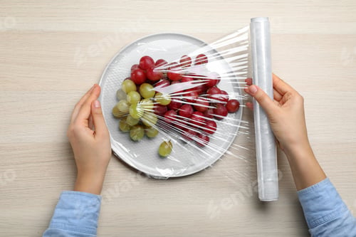Preview: Woman putting plastic food wrap over plate of fresh grapes at wooden table, top view
