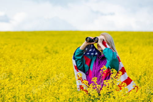 Preview: Woman with USA flag and binoculars in yellow rapeseed field
