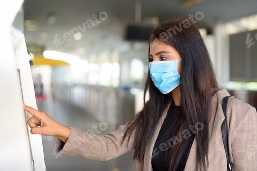 Preview: Profile view of young Indian businesswoman with mask buying ticket at skytrain station
