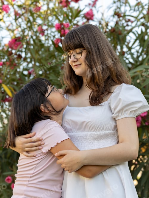 Preview: Teenage girl hugging younger sister in a garden, looking at each other with admiration