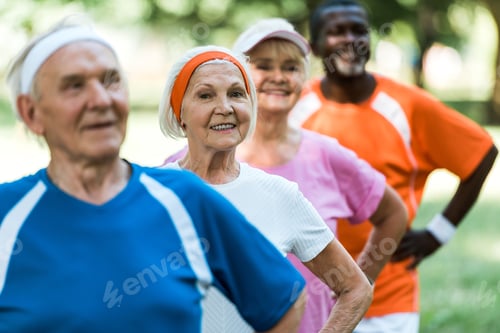 Preview: selective focus of cheerful retired woman standing with hands on hips with multicultural pensioners