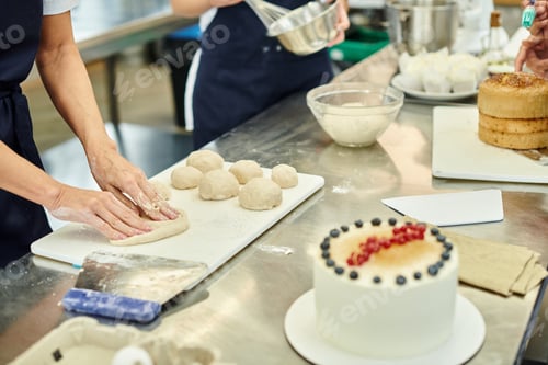 Preview: cropped view of mature chef in apron working on dough next to her young colleague, confectionery
