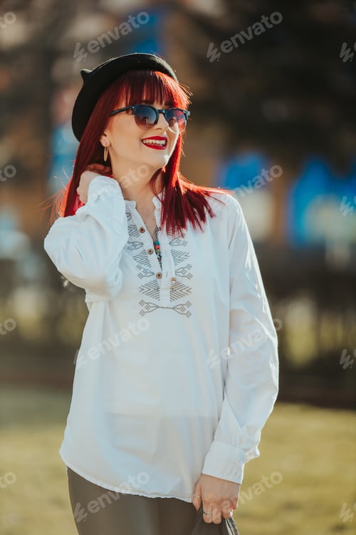 Preview: Vertical shot of a young smiling female with red hair, sunglasses, and a hat posing in a park