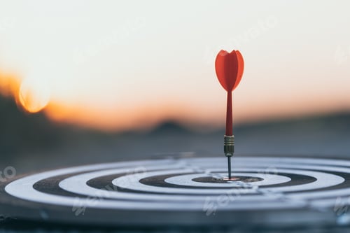 Preview: Close up red dart arrow hitting target center dartboard on sunset background.