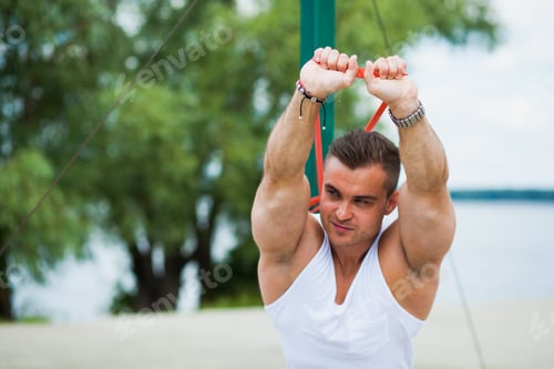Preview: Athletic young man outdoors in summer, hard workout with elastic rubber loop expander.