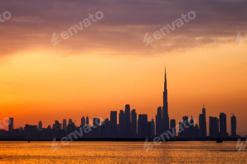 Preview: Silhouette view of Dubai city skyline against the orange dusk sky as seen from the sea at sunset