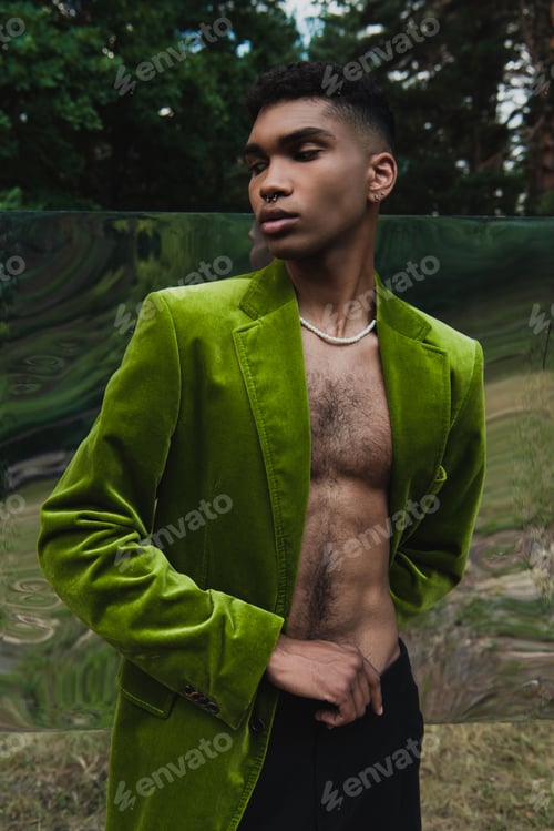 Preview: fashionable african american man in beads and velvet blazer posing in forest near flexible mirror