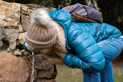 Preview: Mature woman hiking in national park quenches thirst at drinking fountain. Leisure activity