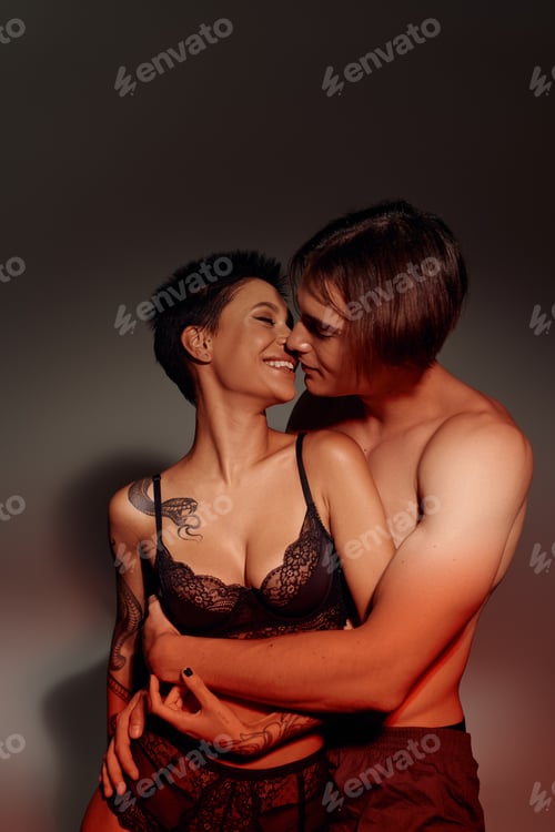 Preview: cheerful and sexy woman in bra kissing with shirtless man embracing her on grey background