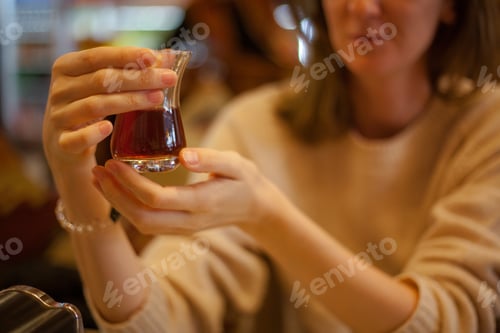 Preview: Blonde woman in a light sweater holding turkish tea glass with tea in her hands