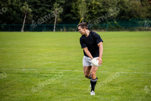 Preview: Male athlete standing on grassy field in sportswear holding white rugby ball by fence, copy space
