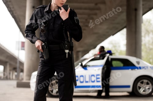 Preview: cropped shot of policewoman using walkie-talkie with blurred partner near car on background