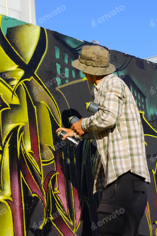 Preview: Rear view of a young graffiti artist spray painting on a concrete wall on the street
