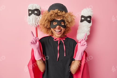 Preview: Cheerful curly woman happy to complete housework makes peace gesture smiles gladfully dressed in sup