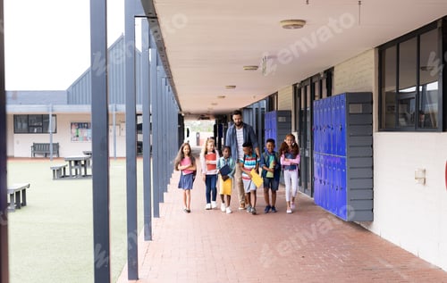 Preview: Diverse, happy male teacher and elementary schoolchildren walking in school corridor, copy space